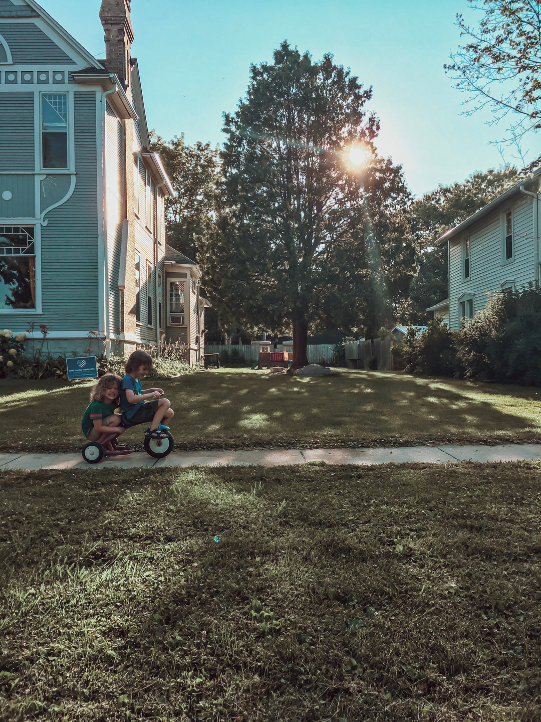 Monica Storey photography.  Children riding on a tricycle on a sidewalk, in front of a home, blue sky, sun shining through the trees.